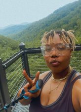 { Black woman with blond locs wearing a black sleeveless top, standing on an overlook platform, holding up the peace sign with the trees of the Pisgah National Forest in the background. The sky is light blue.