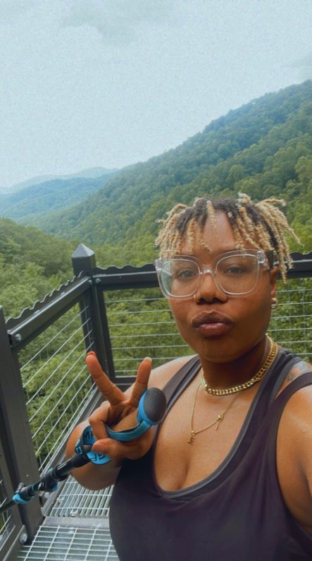 { Black woman with blond locs wearing a black sleeveless top, standing on an overlook platform, holding up the peace sign with the trees of the Pisgah National Forest in the background. The sky is light blue.